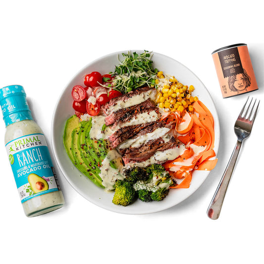 A protein bowl topped with steak, avocado, and veggies, next to a bottle of Primal Kitchen Ranch and Just Spices Avocado Topping.
